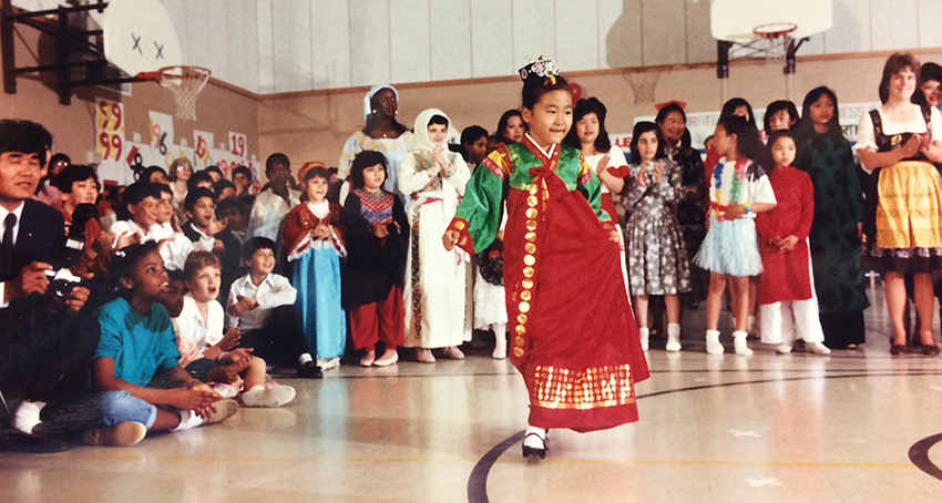 Color photograph from Parklawn Elementary School’s 30th anniversary celebration. Similar to a modern International Festival at Parklawn, the children are dressed in traditional clothing from the country of their native heritage. A girl in a red and green dress parades before a diverse crowd of students and parents gathered in the school’s gymnasium. 